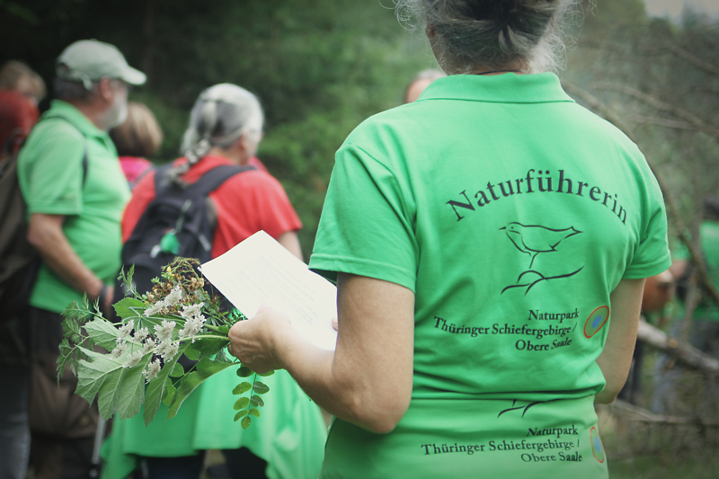 Eine Gruppe Personen ist im Wald zu sehen. Die Frau im Vordergrund tr&auml;gt ein T-Shirt mit der Aufschrift "Naturf&uuml;hrerin".