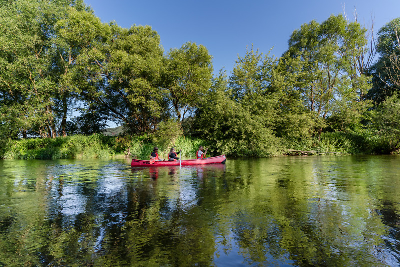 Drei Personen fahren ein rotes Kanu auf einem ruhig flie&szlig;enden Fluss, entlang gr&uuml;n bewachsener Auen.
