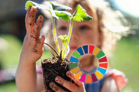 Kleines M&auml;dchen h&auml;lt ein junges Pfl&auml;nzchen in den H&auml;nden. Au&szlig;erdem im Bild ist ein bunter Kreis mit den Farben der 17 Ziele f&uuml;r Nachhaltigkeit.
